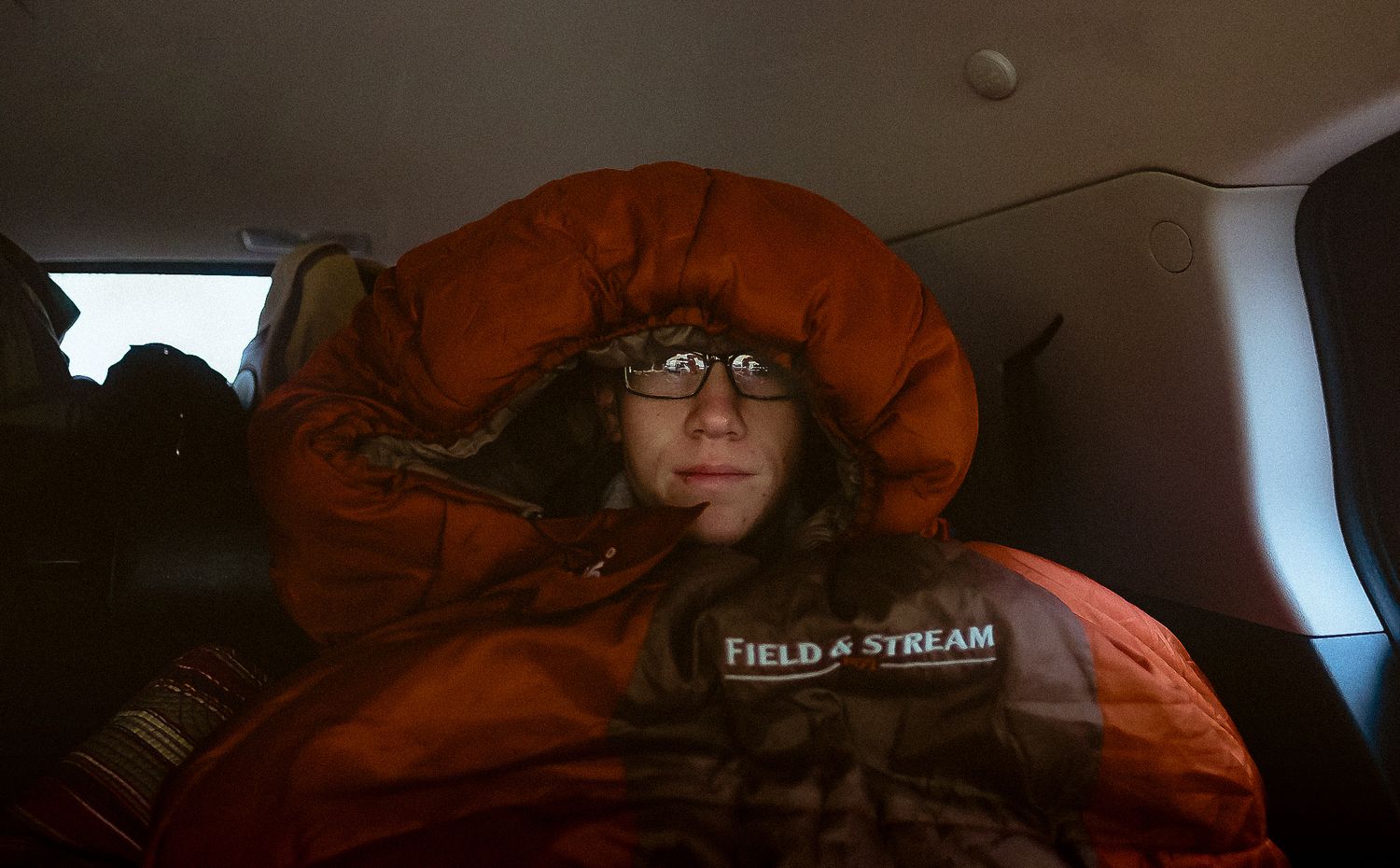 Person wrapped in red sleeping bag wearing glasses relaxing in car backseat.
