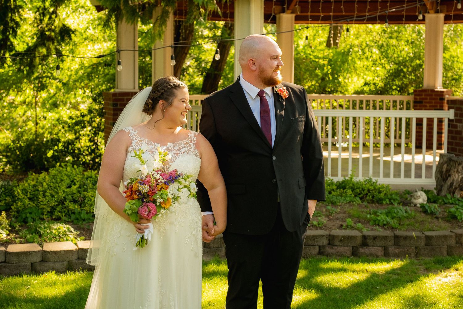 A wedding couple poses together in a garden setting with white gazebo and floral bouquet.