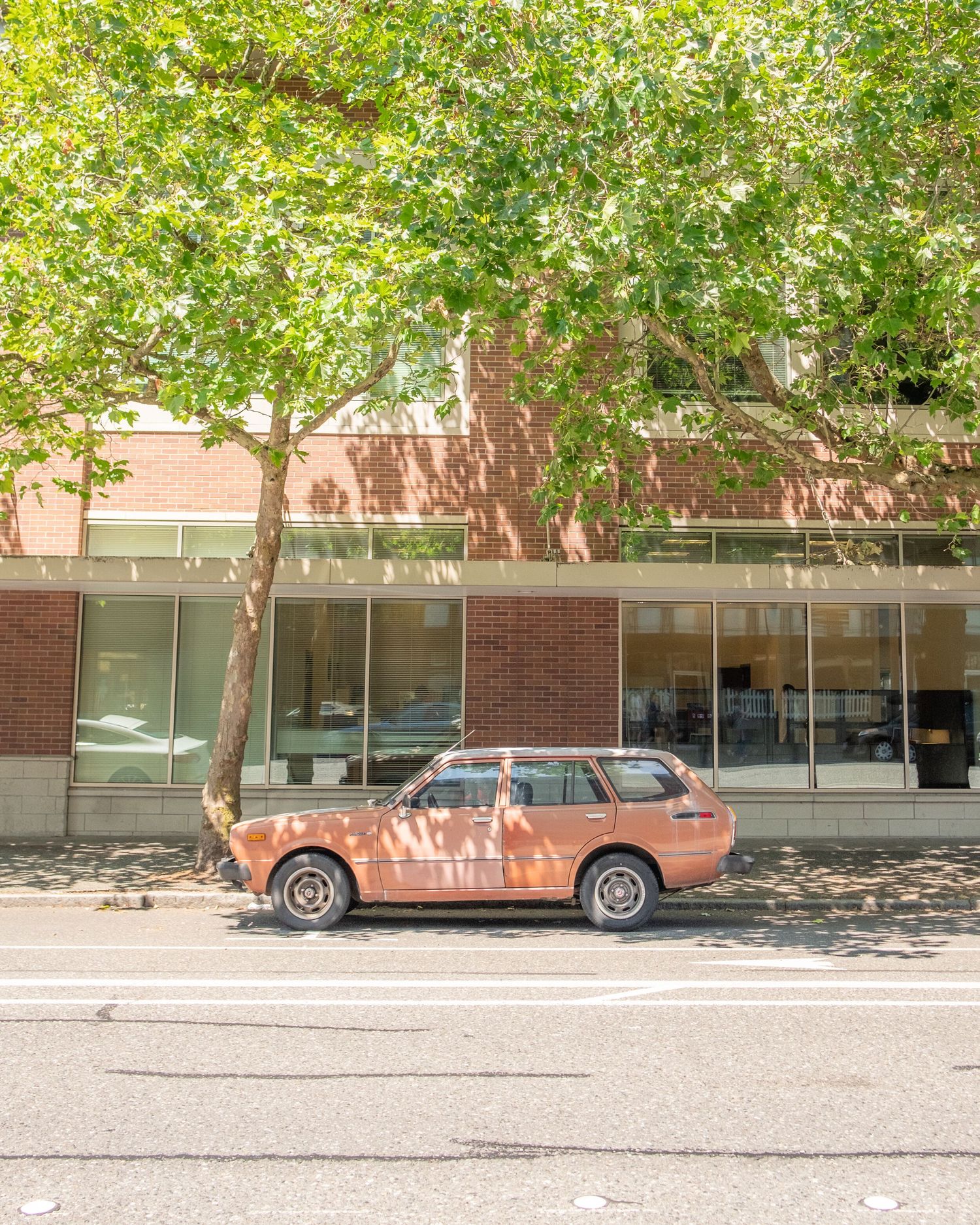 Vintage brown car parked outside brick building with green trees providing shade.