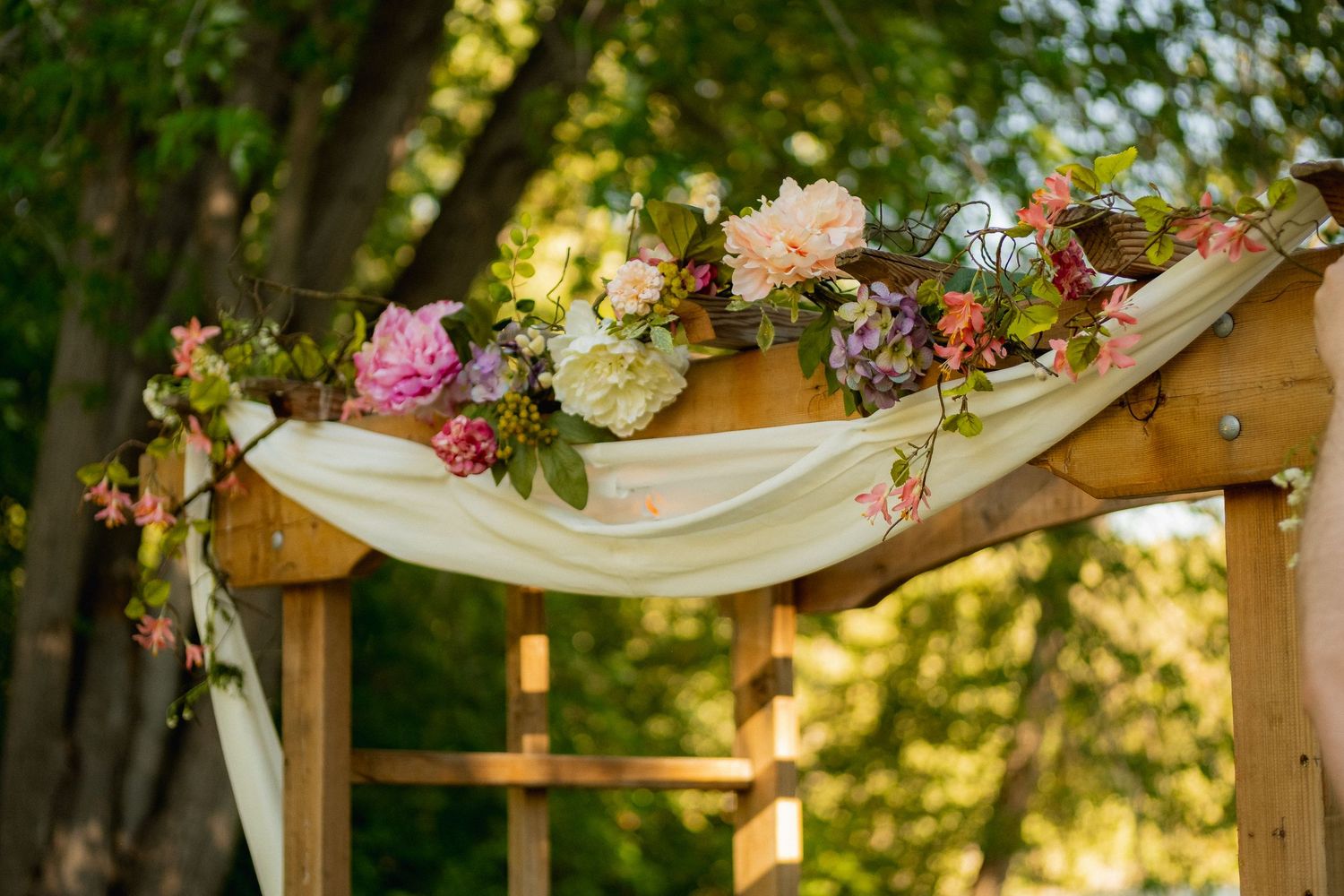 A wooden wedding arch decorated with colorful flowers and draped white fabric in a garden setting.