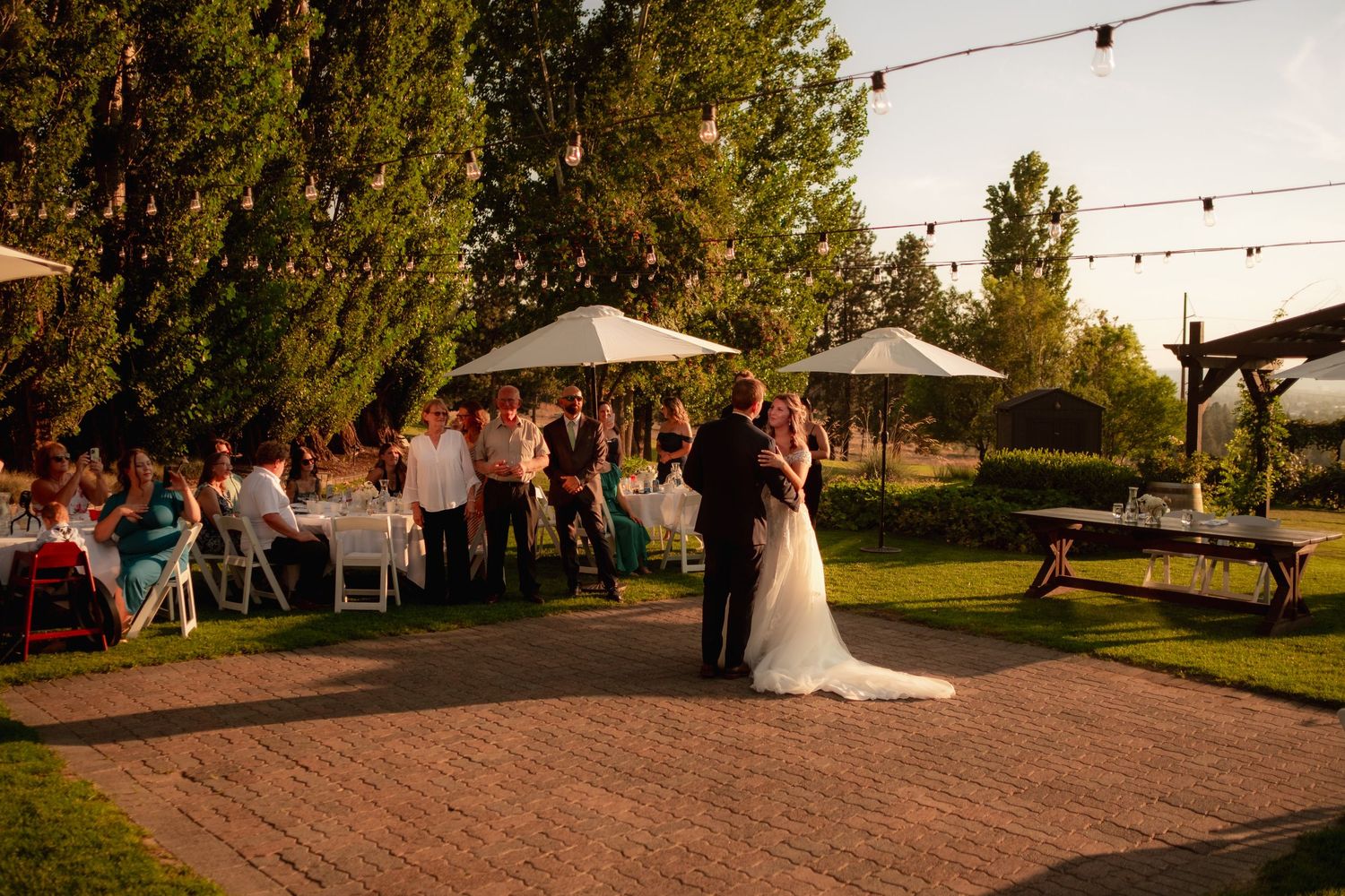 Outdoor wedding reception setup with string lights and a paved courtyard at sunset.
