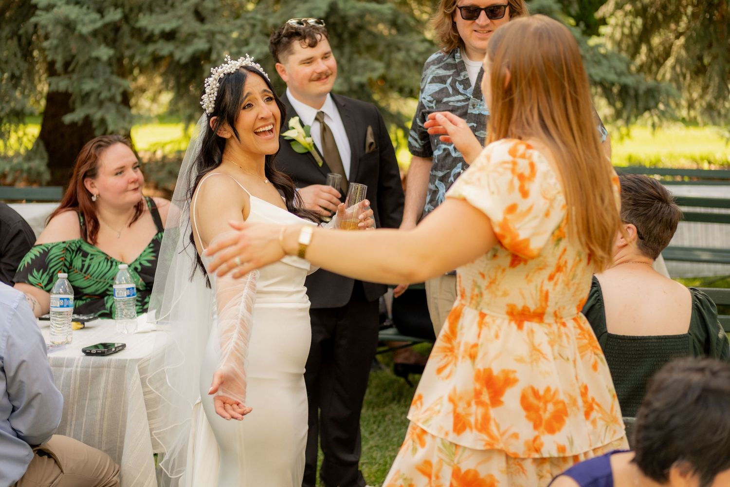 Bride in white gown and veil laughing with guest in orange dress at outdoor wedding reception.