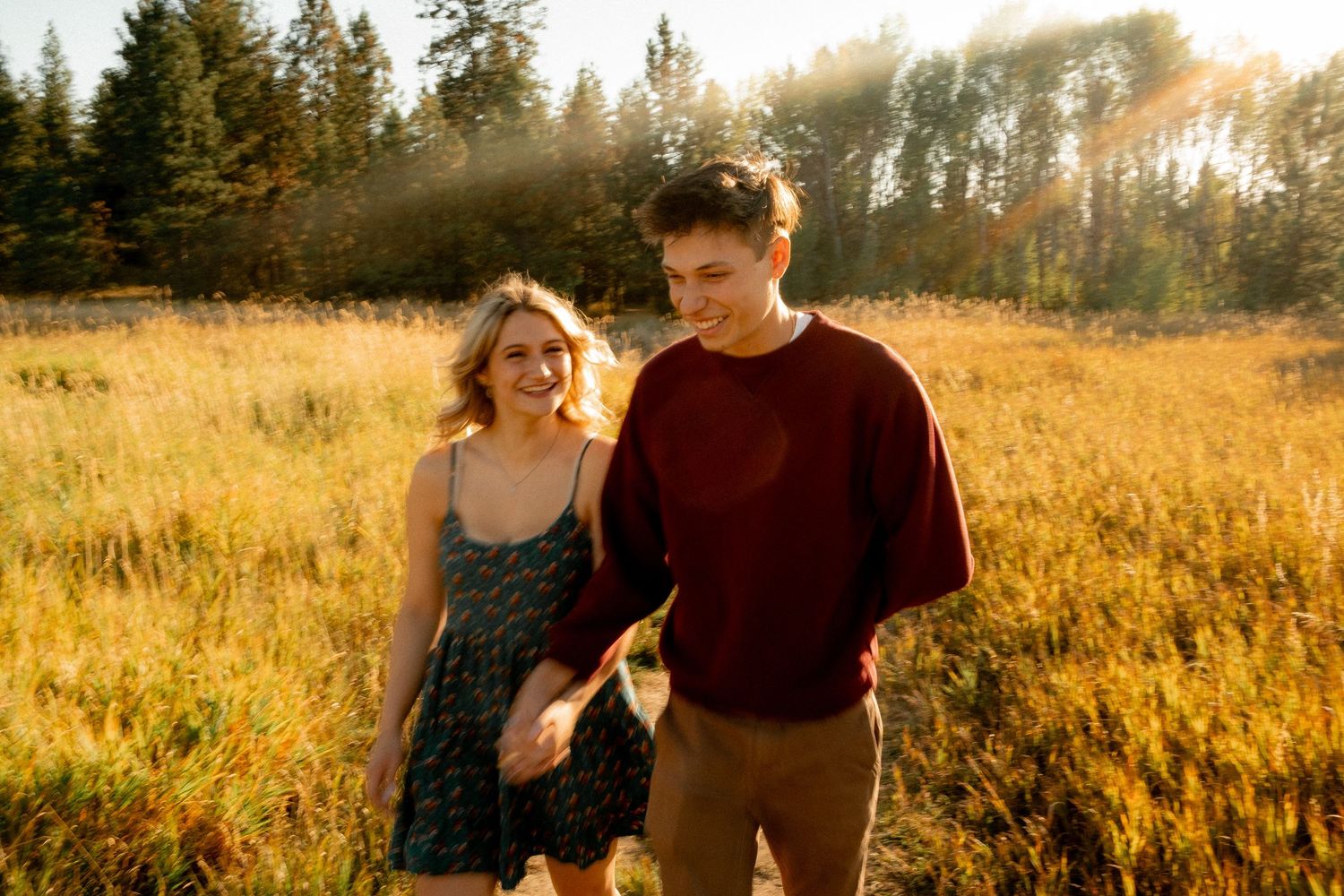 People walking through a scenic mountain meadow with sunlit evergreen trees in the background.