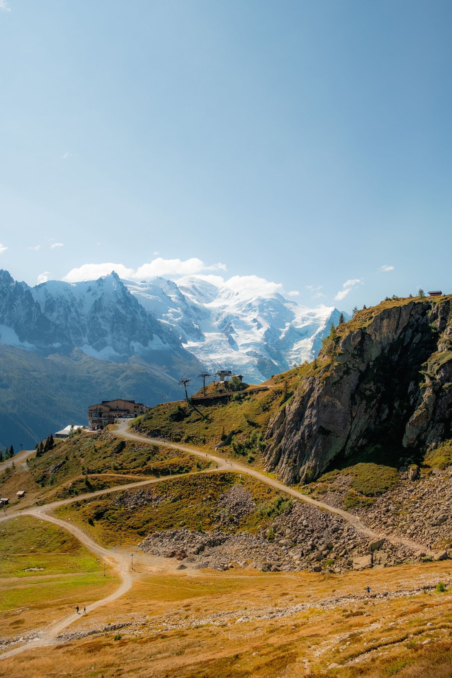 Winding mountain path leading through alpine terrain with snow-capped peaks and rugged cliffs in the background.