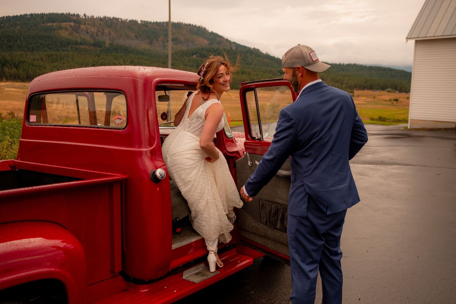 A classic red vintage pickup truck parked near a rural building with two people in formal attire.