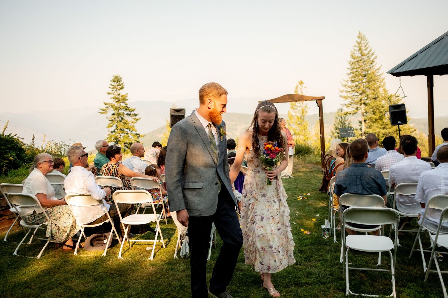 Wedding guests gather outdoors at sunset on a hilltop venue with tables and chairs arranged for a celebration.