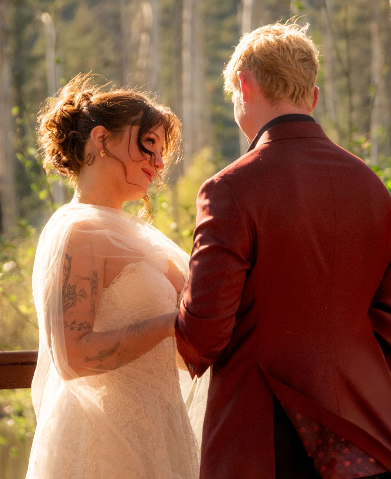 A bride in a white lace gown and groom in a burgundy suit stand together during an outdoor wedding ceremony.
