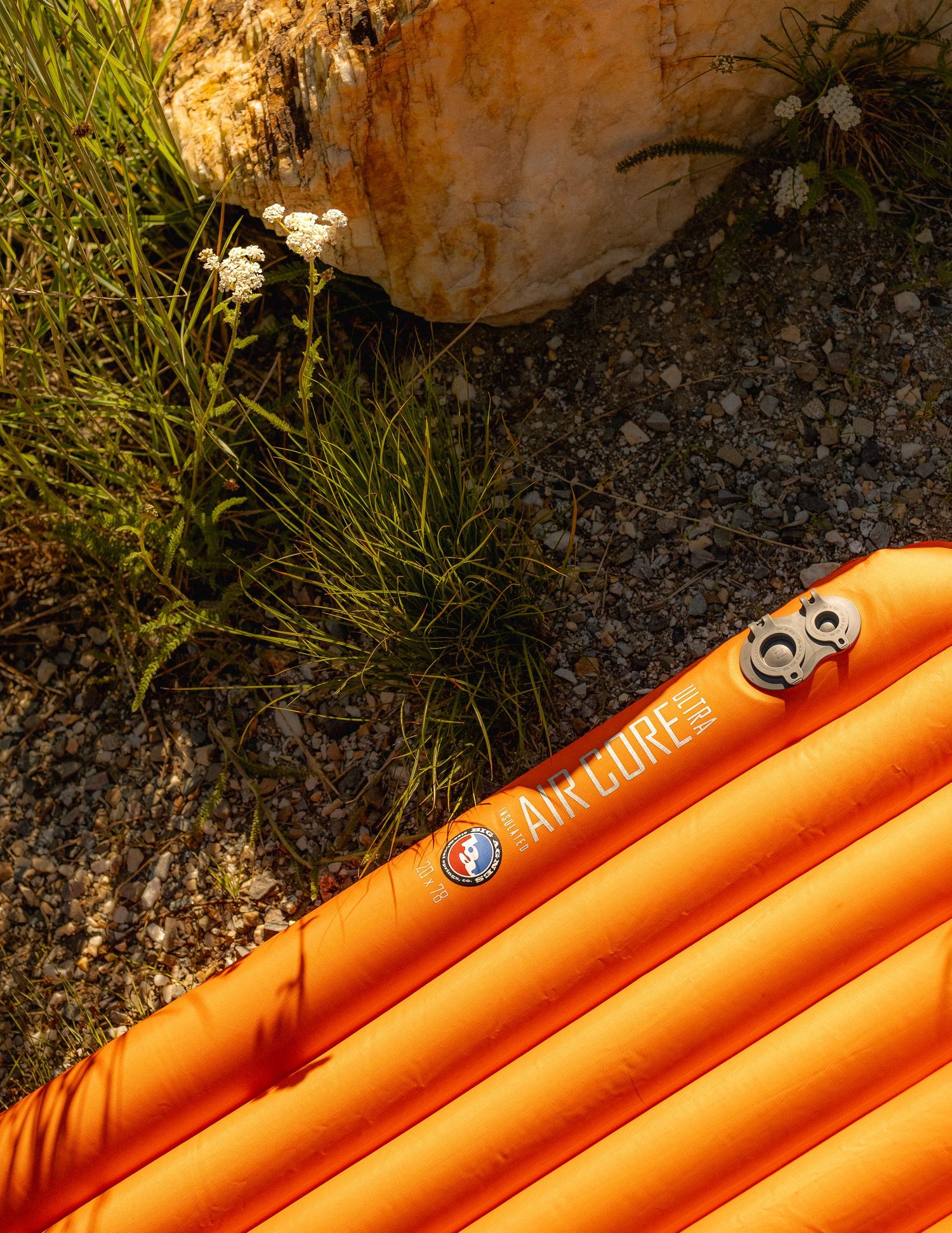 Orange inflatable sleeping pad or camping mat next to white wildflowers against rocky ground.