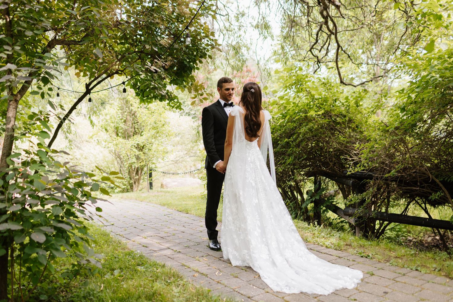 A couple poses on a garden path surrounded by lush greenery and trees.
