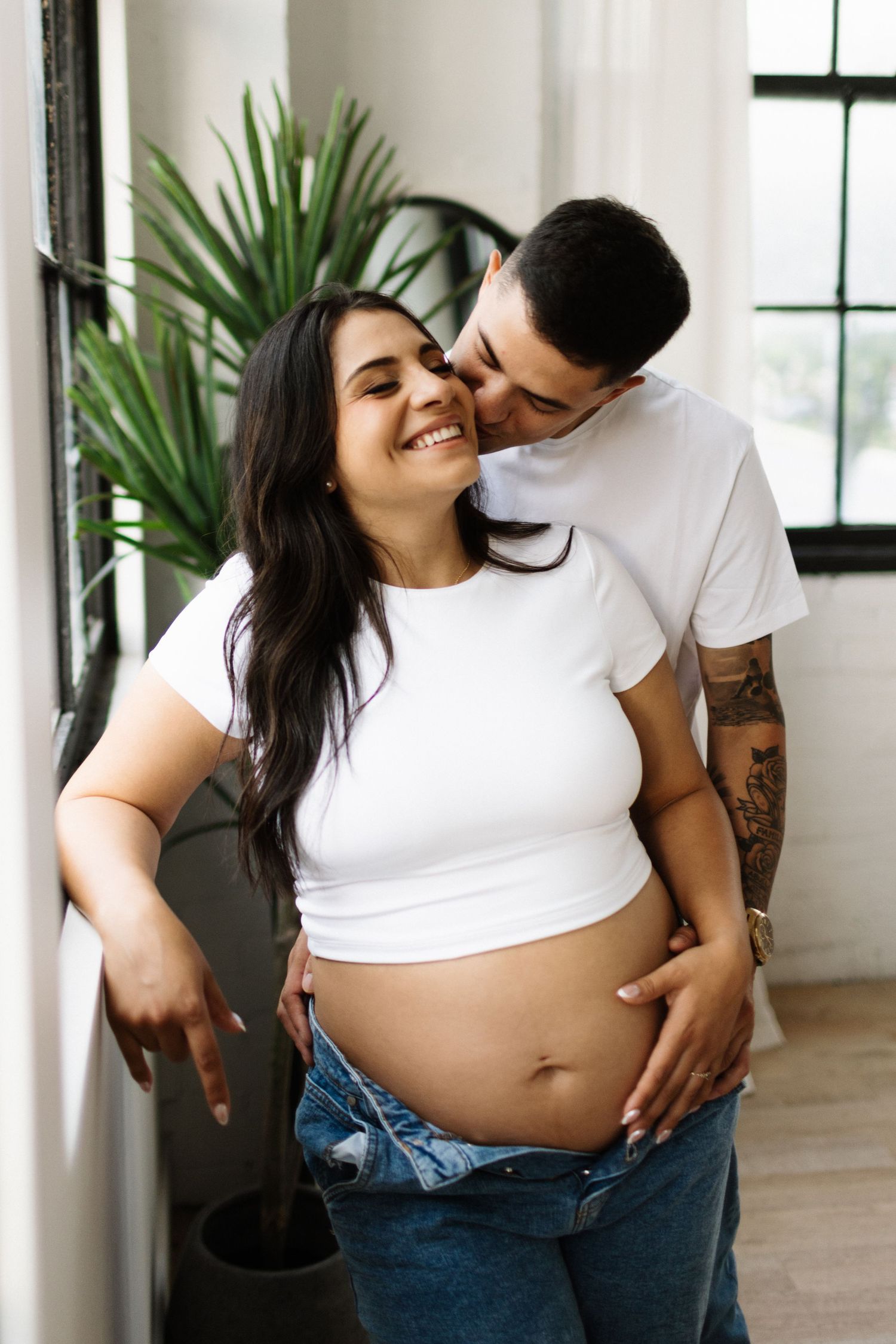 An expecting couple in white shirts share an intimate moment near a house plant, showing off a baby bump.