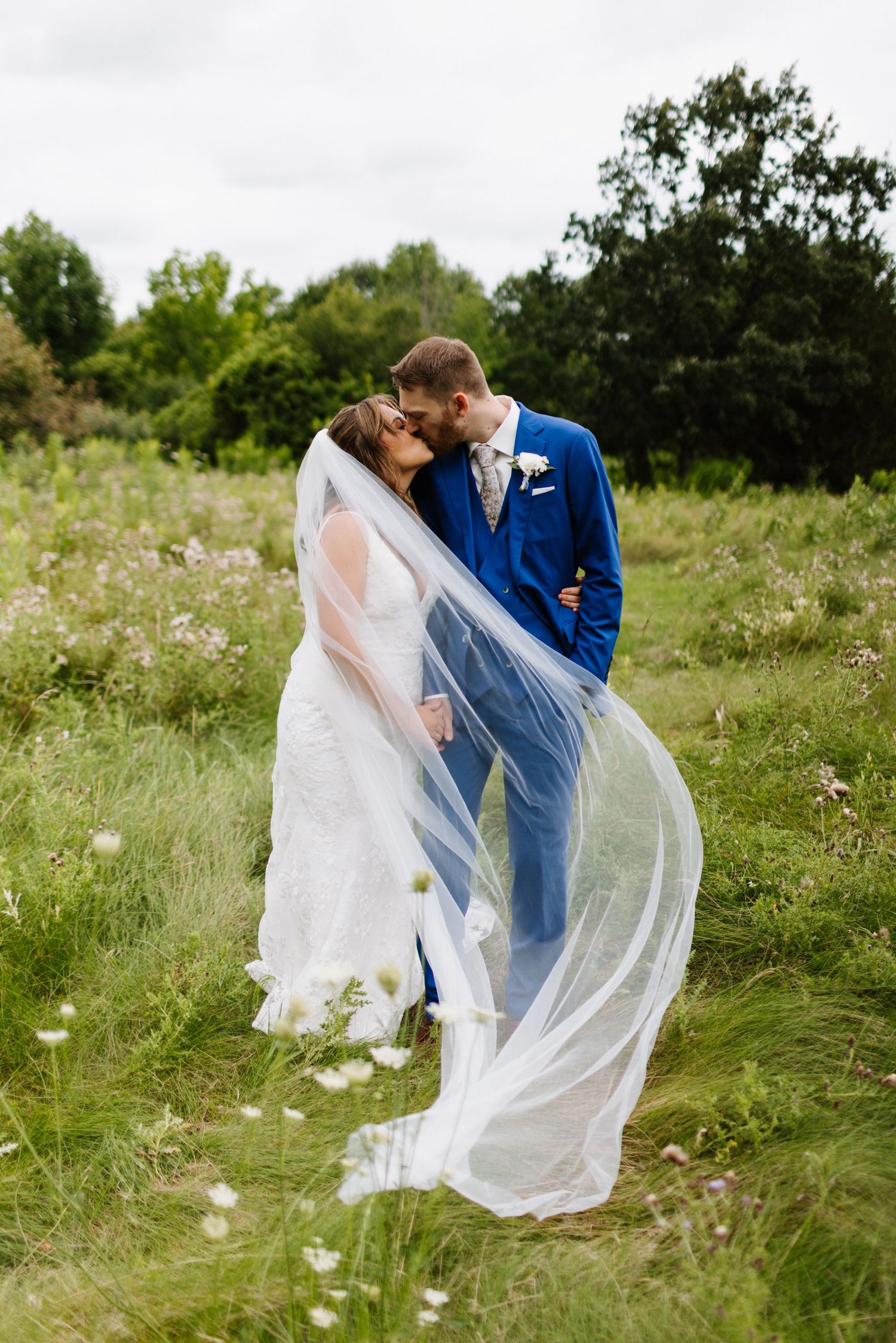 A romantic couple embraces in a wildflower meadow as the bride's long veil flows gracefully in the breeze.