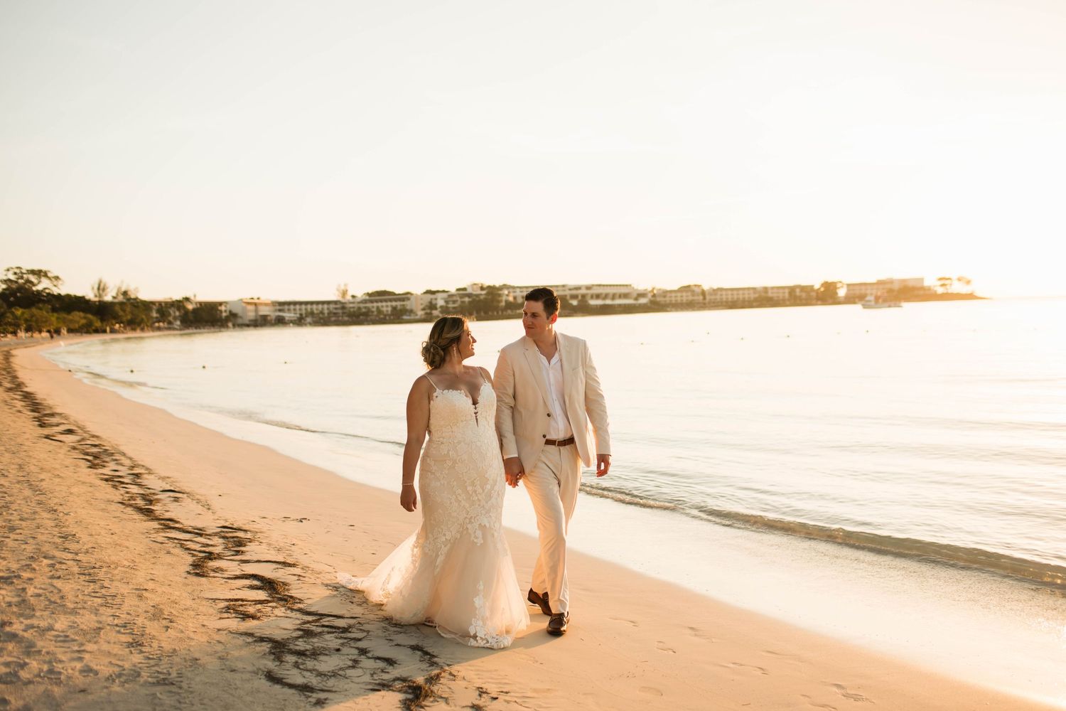 A couple walks along a sandy beach at sunset in white wedding attire.