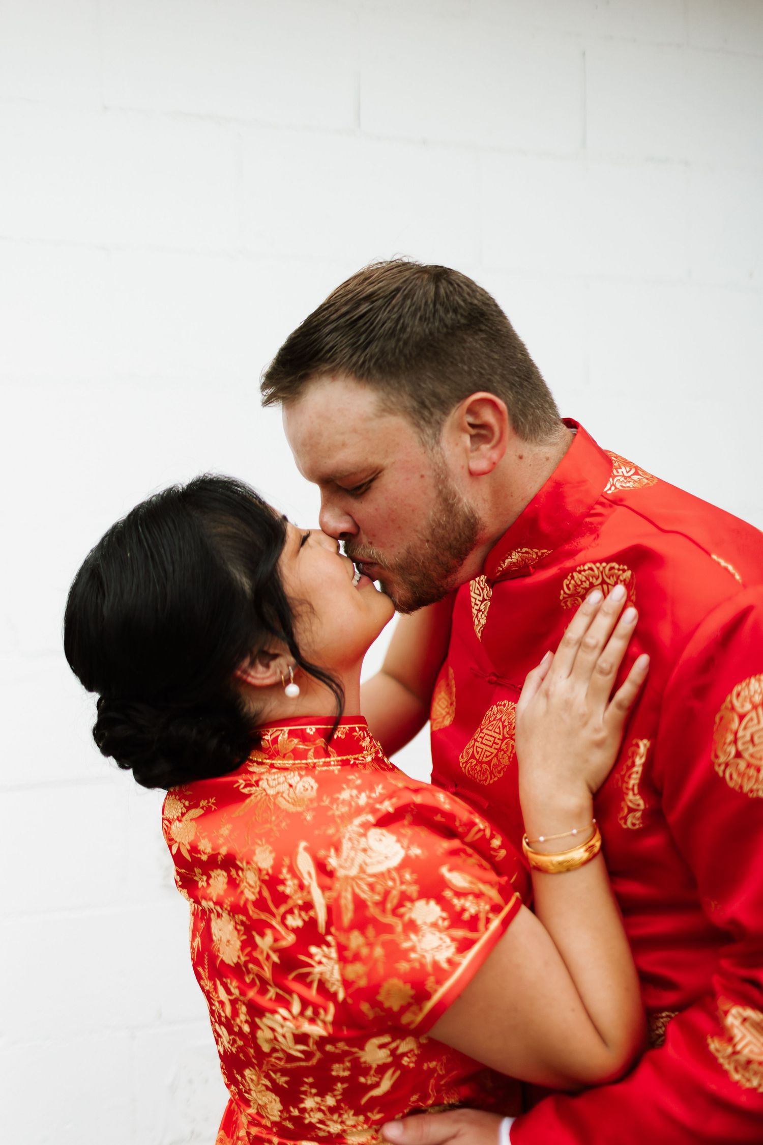 A couple in traditional red Chinese wedding attire share a romantic kiss against a white wall.