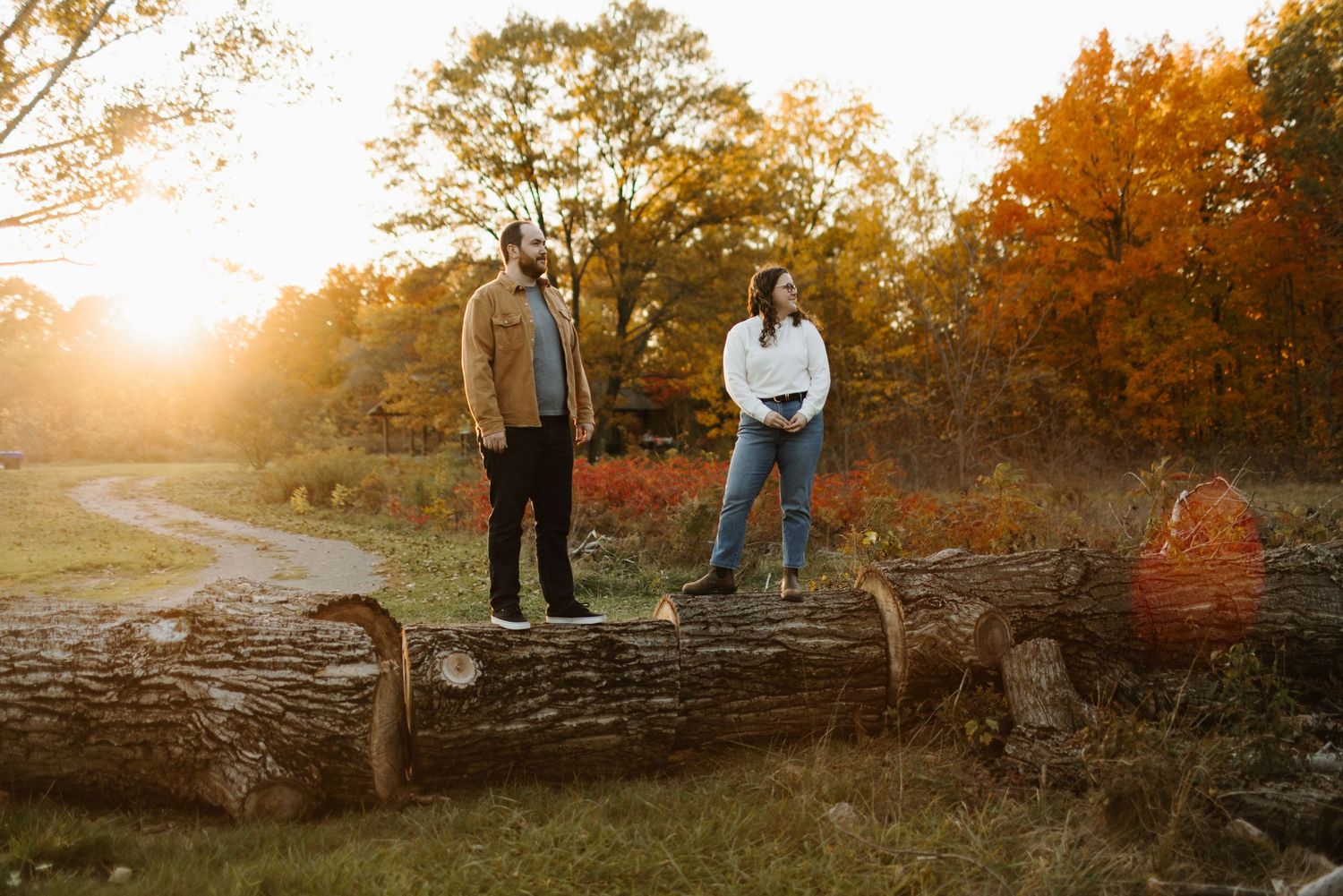 Two people pose on fallen tree trunk at sunset with autumn foliage and golden light in background in Oakville Ontario