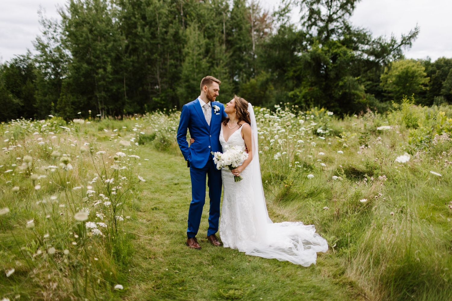 A couple stands together in a wildflower meadow during their wedding, with a blue suit and white dress.