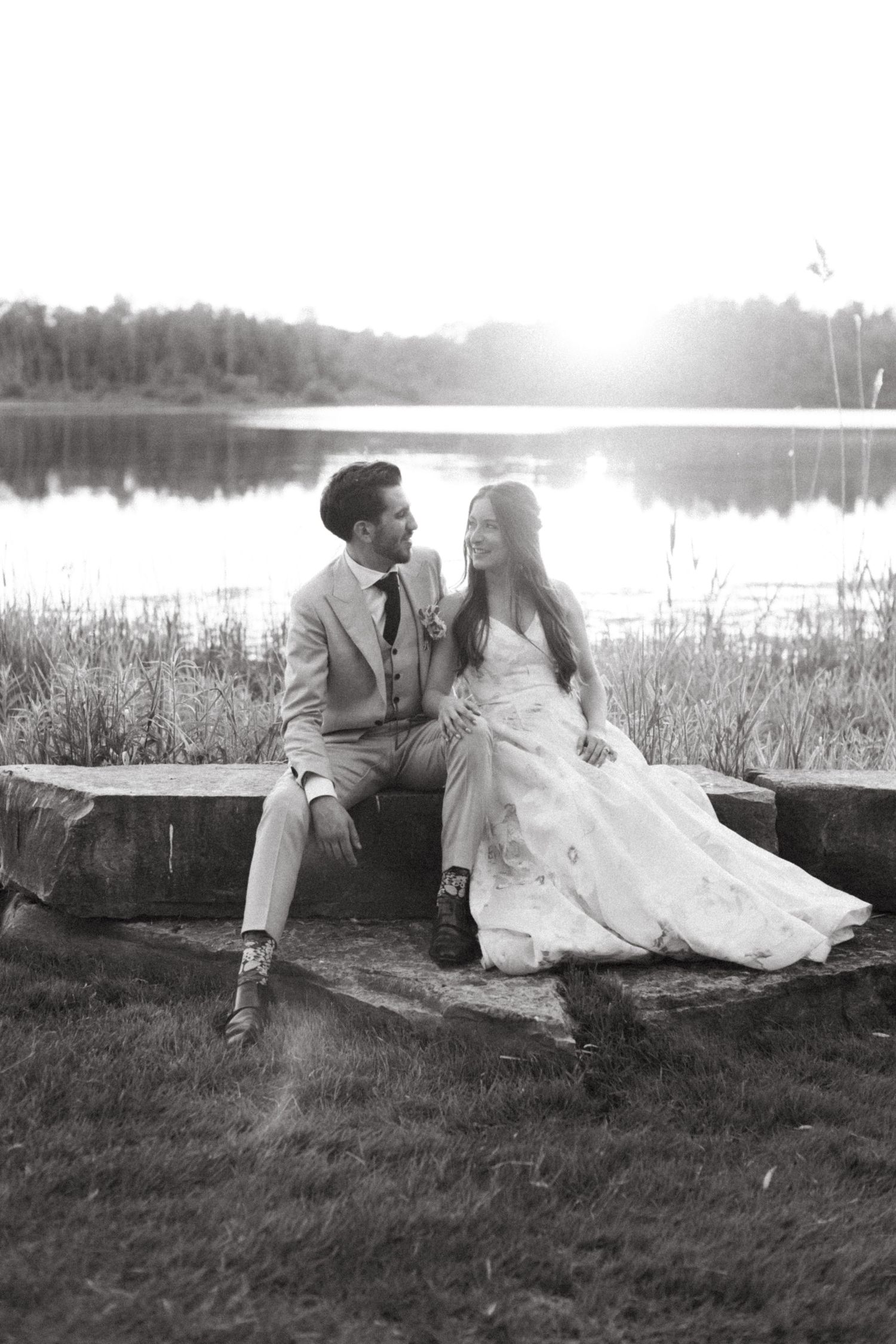 A couple in wedding attire sits together by a serene lake at sunset in black and white.