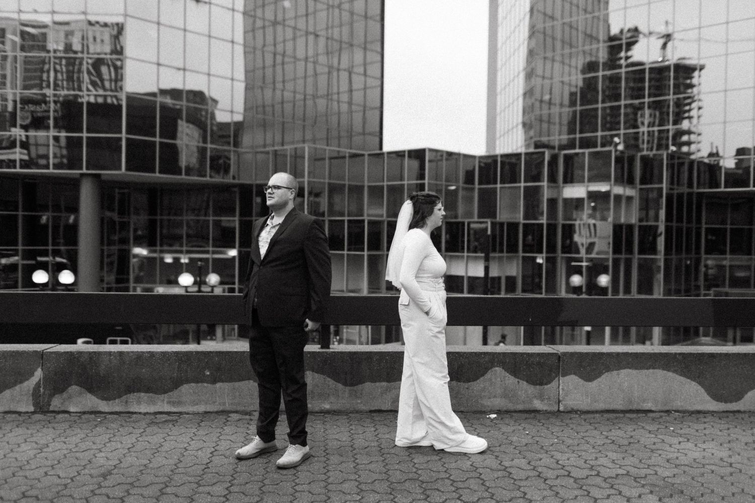 Black and white portrait of couple standing against reflective glass building exterior.