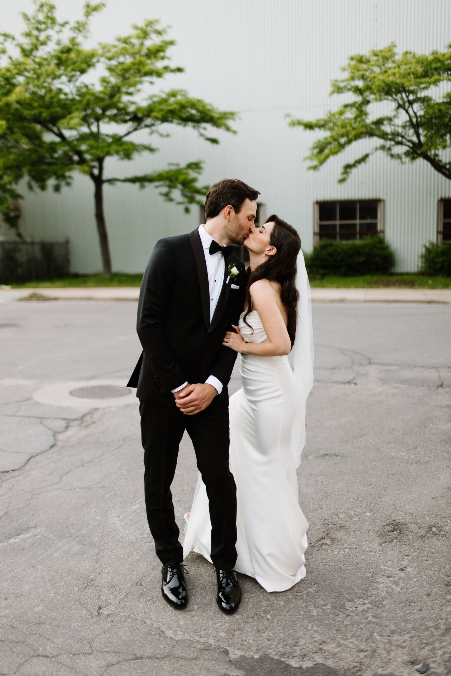 A romantic wedding couple shares a kiss outdoors, the groom in classic black tuxedo and bride in a white gown. Taken in Hamilton Ontario