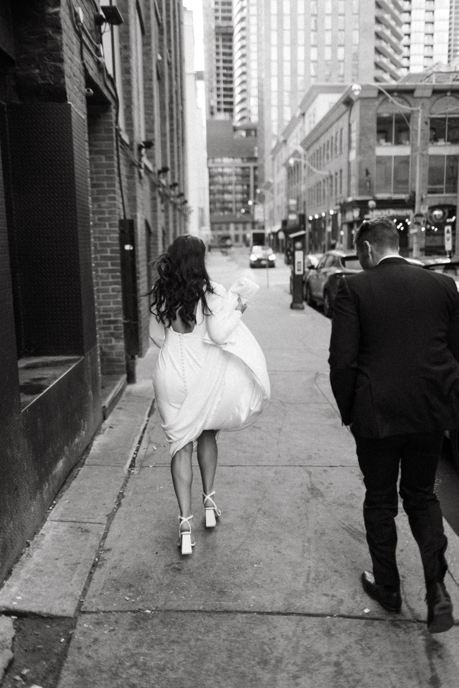A couple walking down a city sidewalk with tall buildings in the background.