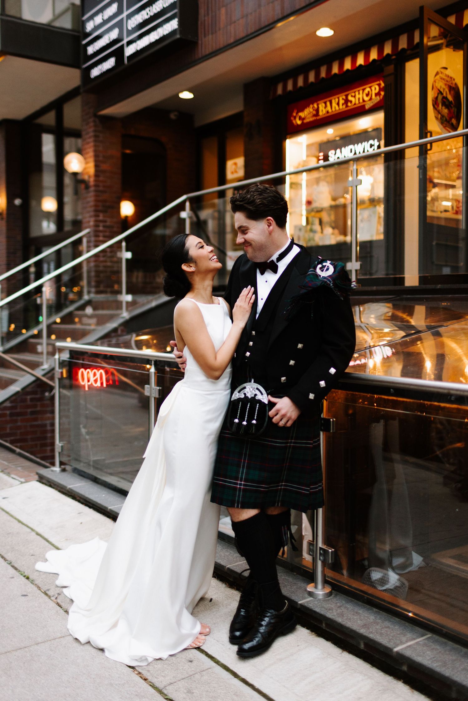 A couple in wedding attire stands outside a bakery shop, with the groom wearing a traditional Scottish kilt at night in Toronto.