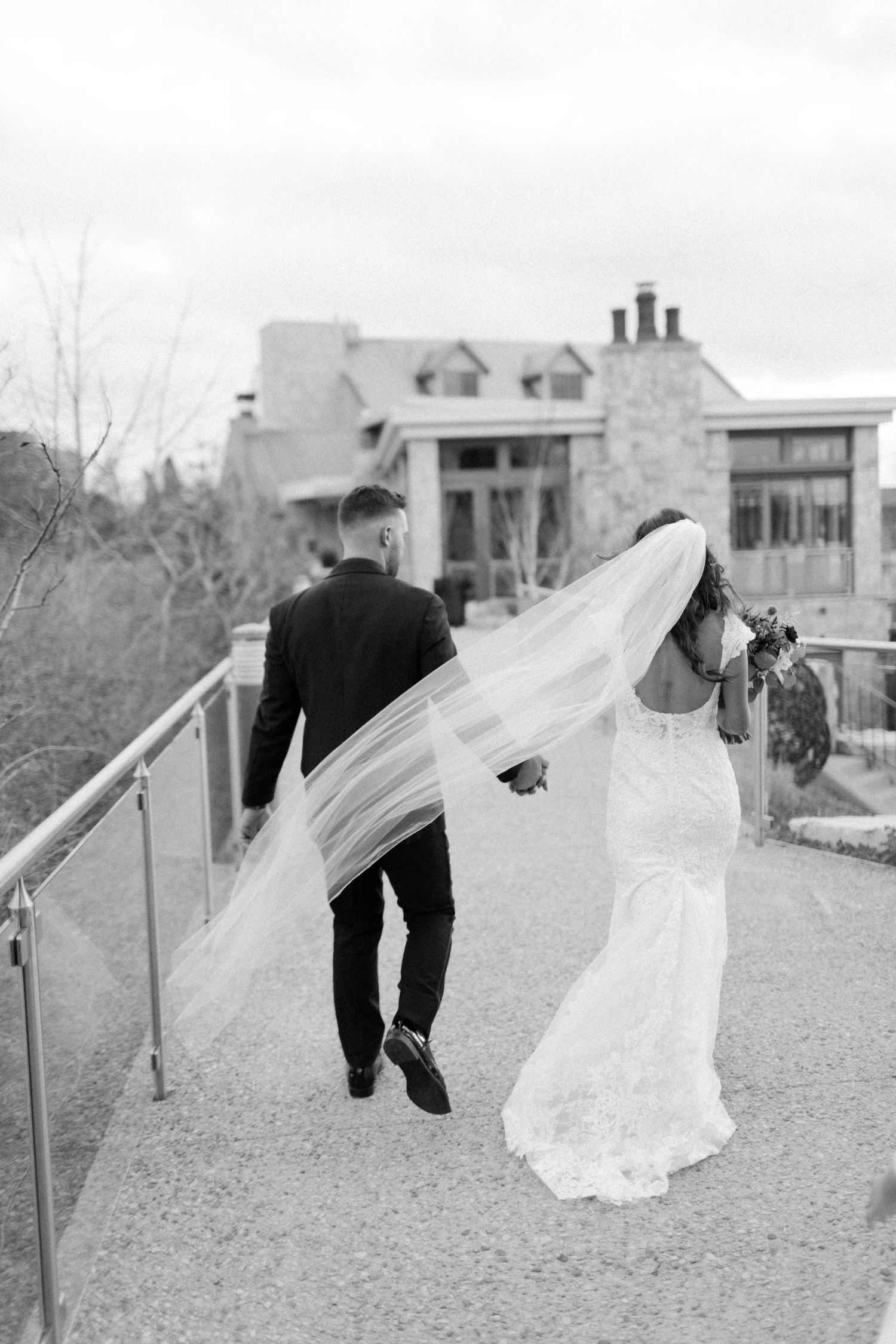 Bride's flowing veil catches the wind as couple walks along pathway near stone building.