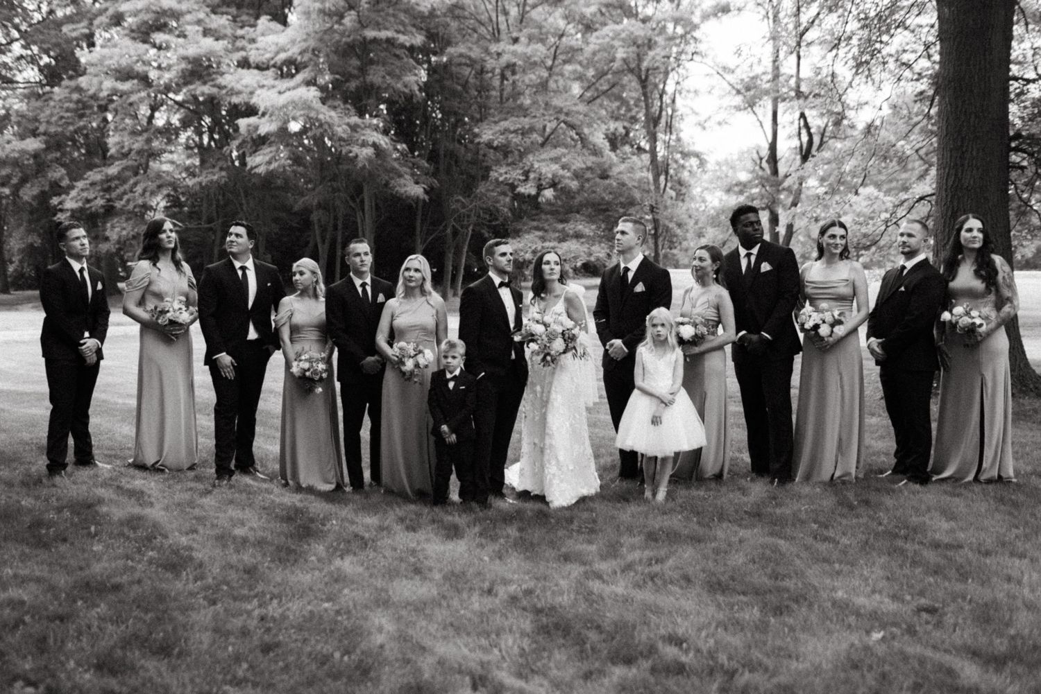 Large wedding party lines up for formal portrait on grass with trees in background.