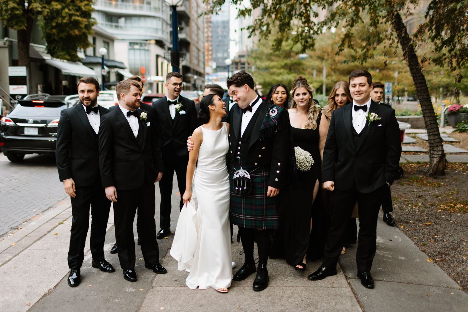 A wedding party in formal attire poses on a city sidewalk.