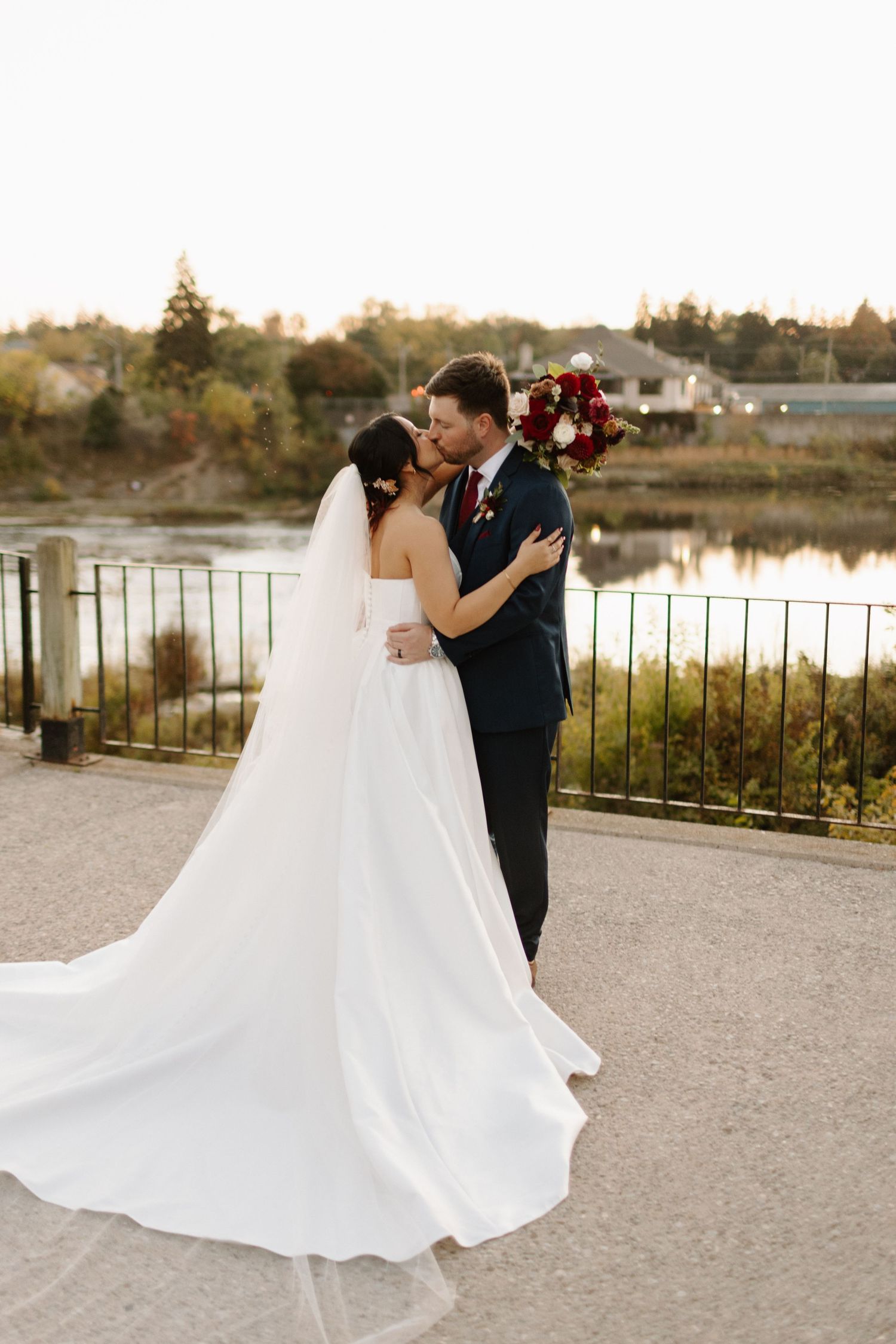 A romantic couple in wedding attire dance and embrace by a waterfront during golden hour.