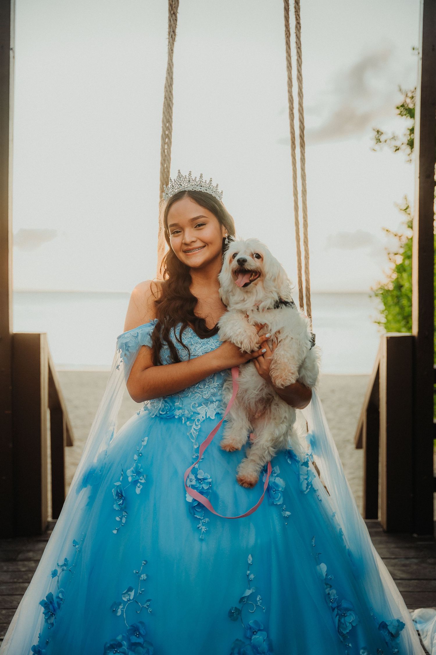 A young person in a blue ballgown poses on a porch swing with a small white dog.
