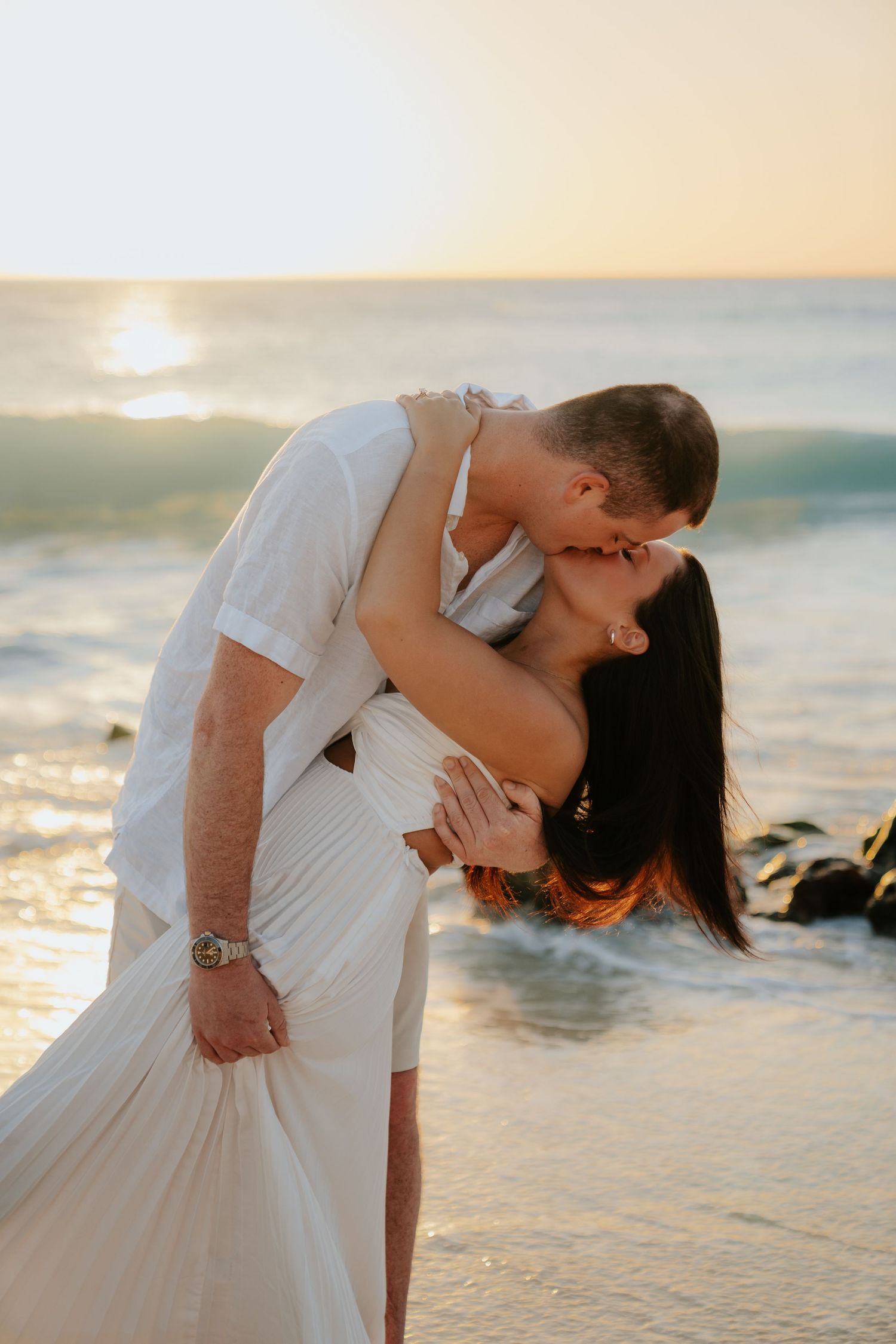 Couple sharing a romantic kiss on the beach at sunset, dressed in white, with ocean waves in the background.