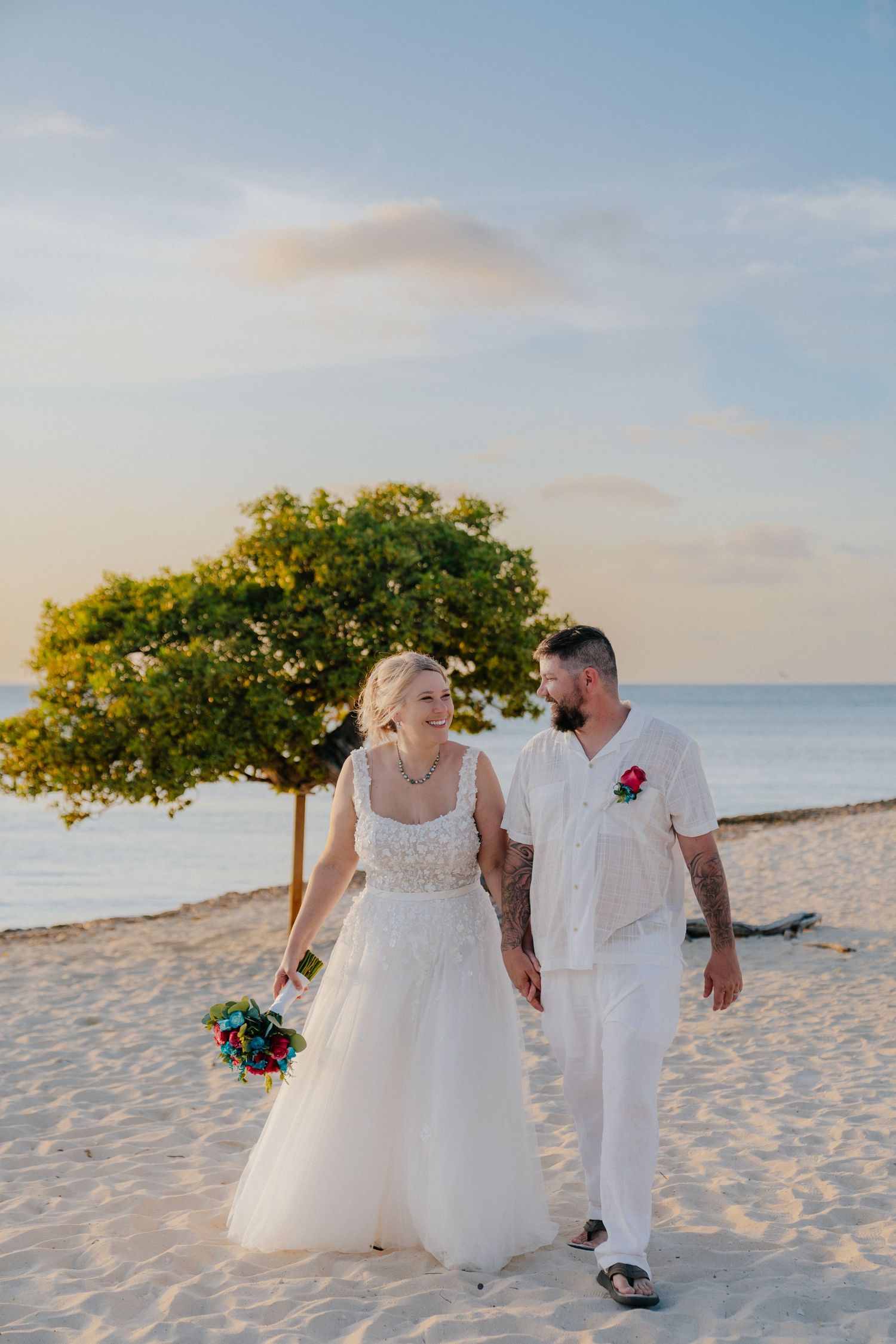 Bride and groom walking hand in hand on a sandy beach at sunset with a tropical tree and ocean backdrop.