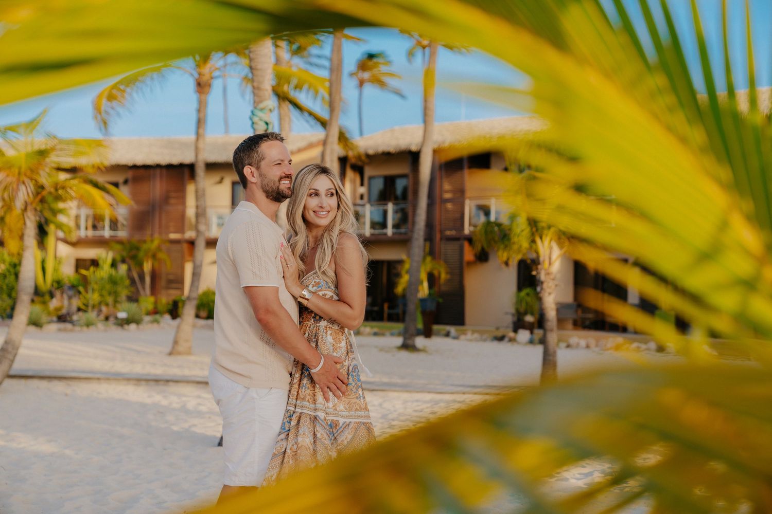 Palm fronds frame a tropical resort setting with colorful buildings and swaying trees.