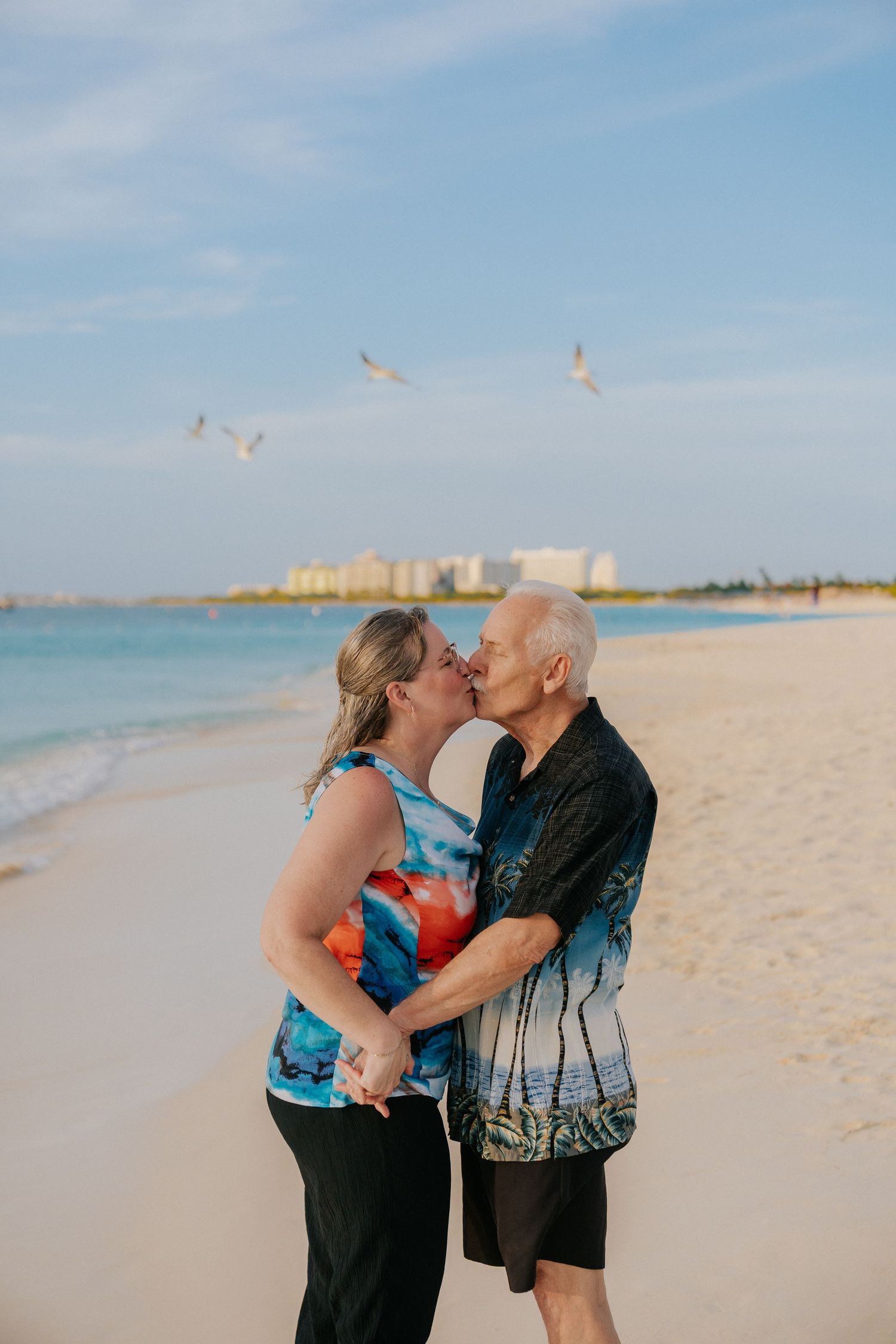 A romantic couple embraces on a sunny beach with seabirds flying in the background.