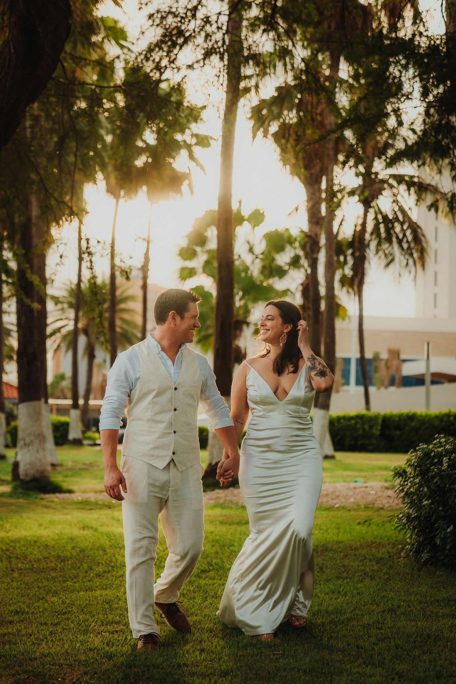 Couple in white attire walking and laughing together in a park with Spanish moss trees.