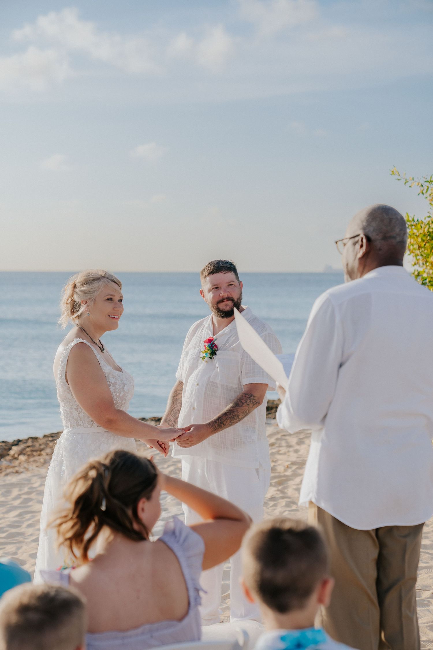 A beach wedding ceremony taking place at sunset with guests seated on white chairs by the ocean.