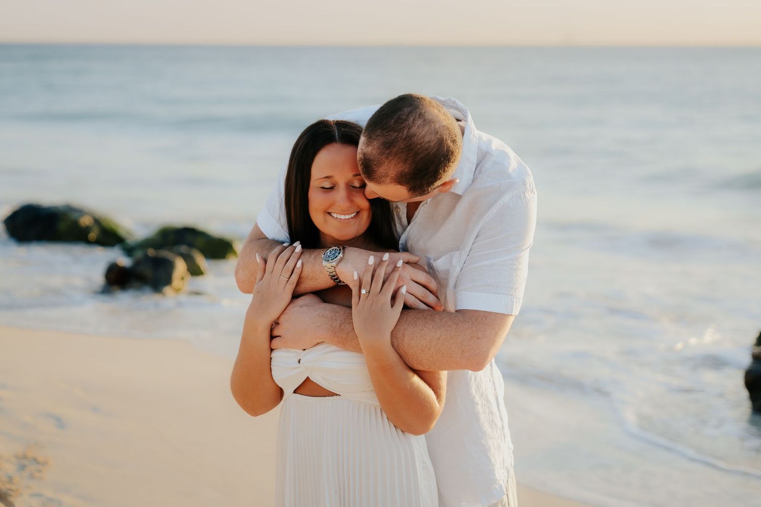 A couple in white outfits embraces lovingly on a scenic beach during sunset.