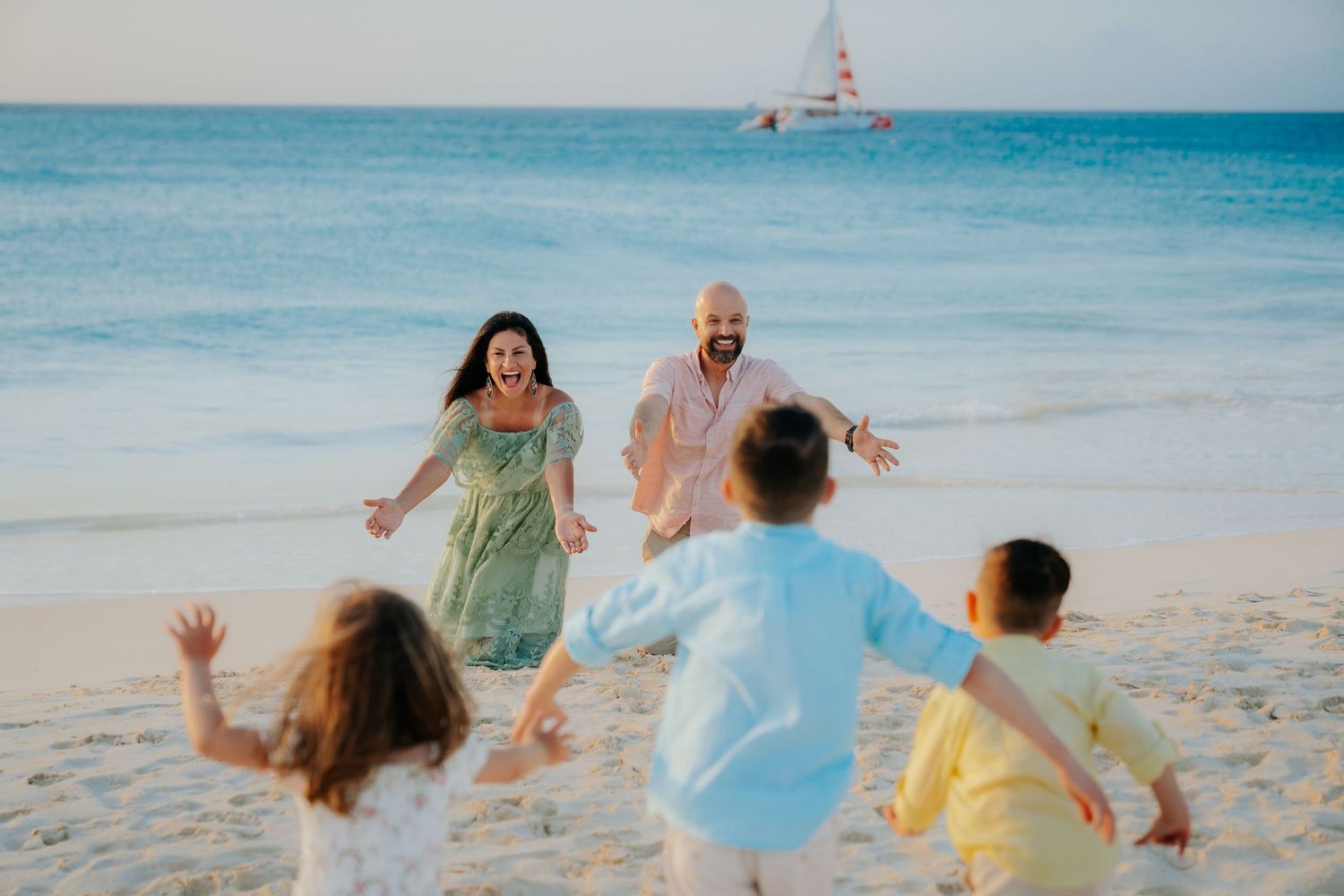 A family runs and plays together on a tropical beach with turquoise waters and a sailboat in the background.