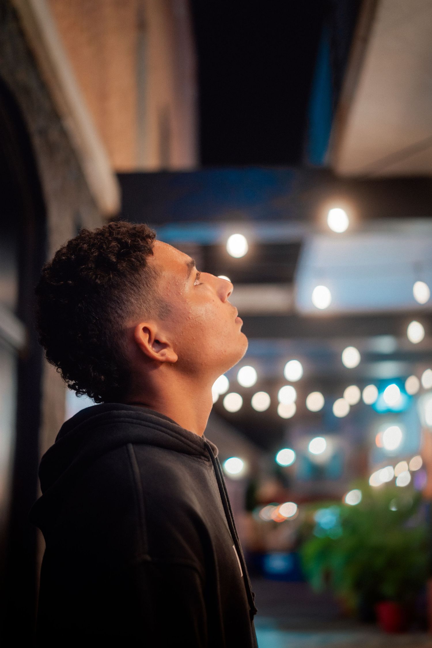 Person in black hoodie gazing upward at string lights in evening setting.