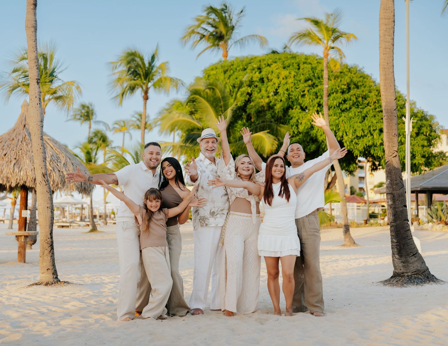 Wedding party in white dresses poses along a tropical beach with palm trees and sand at sunset.