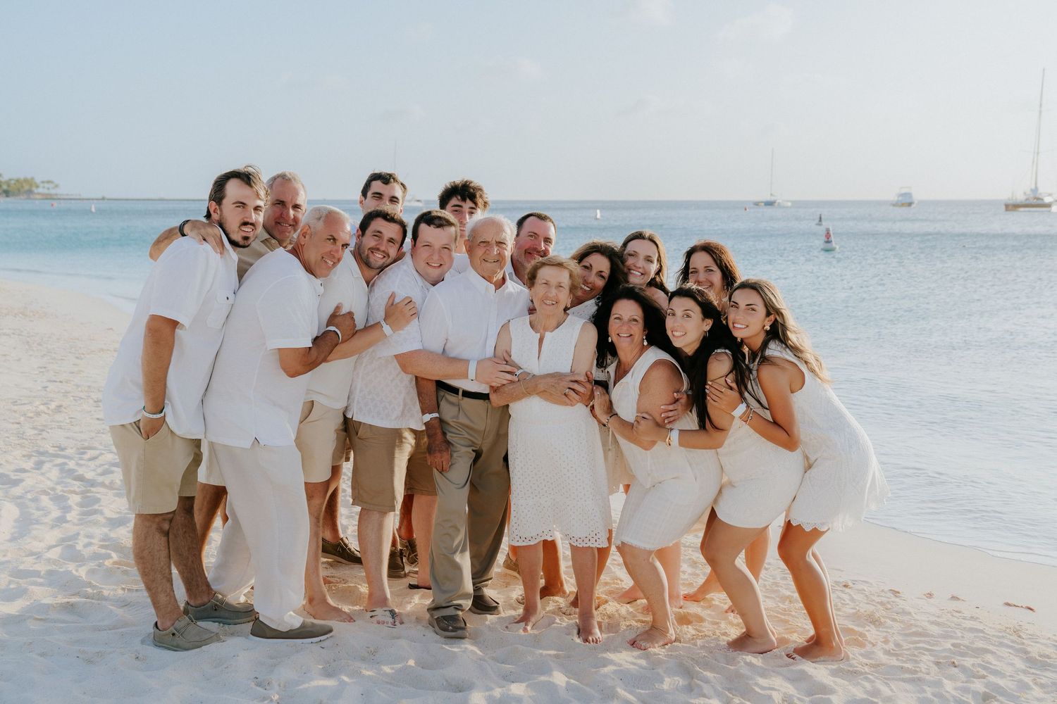 A group in coordinating white outfits poses together on a tropical beach with turquoise water and boats in background.