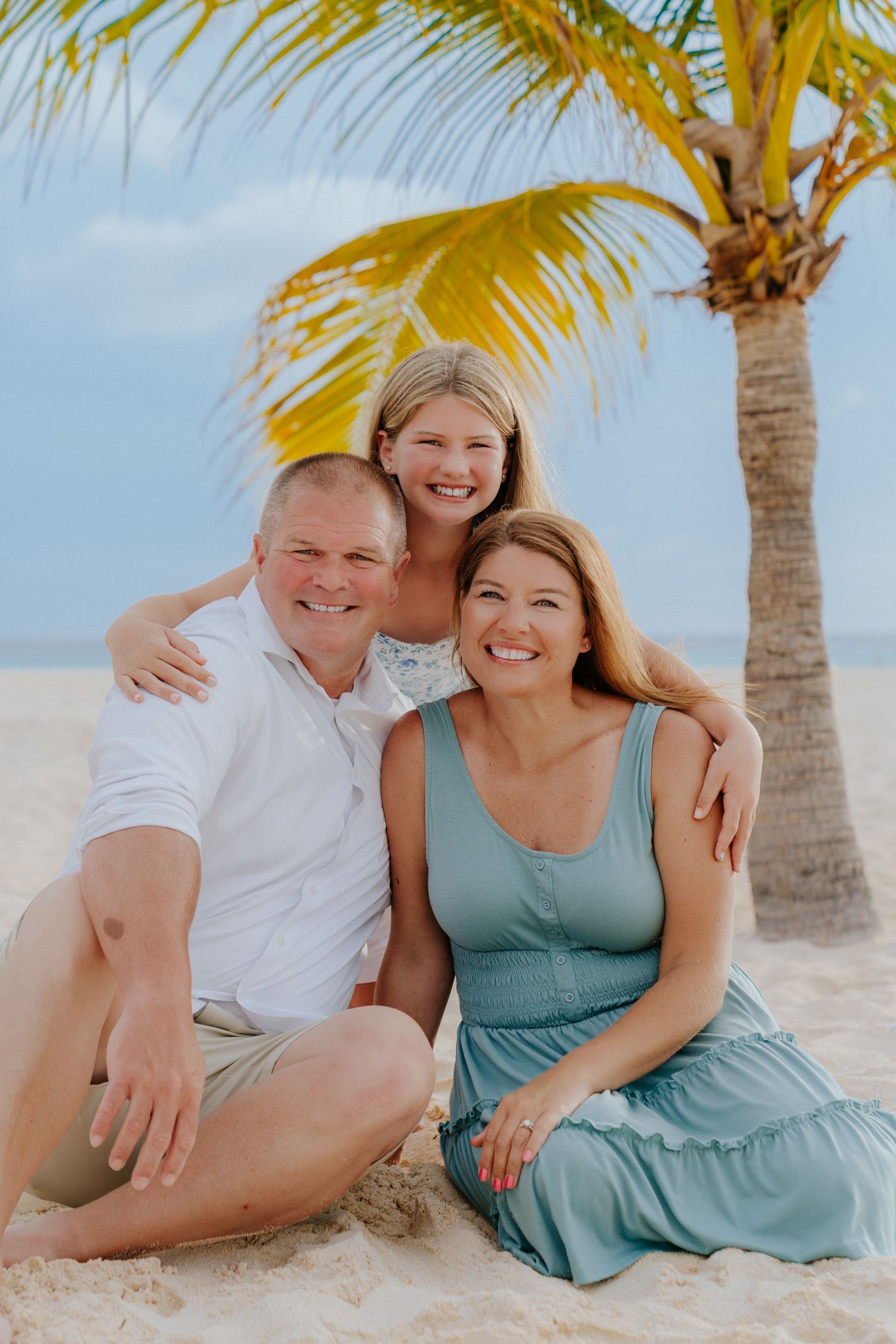 Three people pose together under a palm tree on a sunny tropical beach.