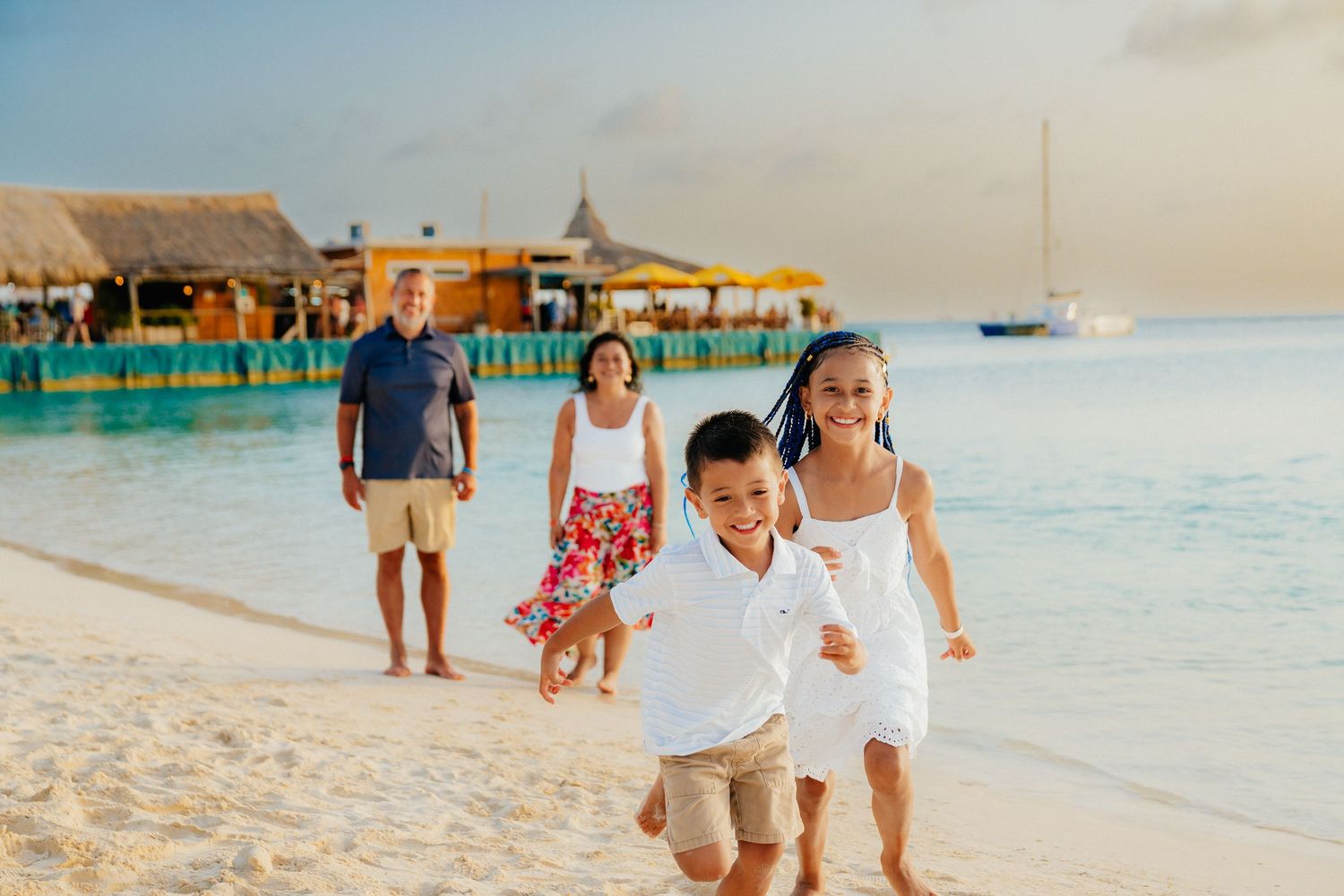 A family walks along a tropical beach with overwater bungalows in the background.