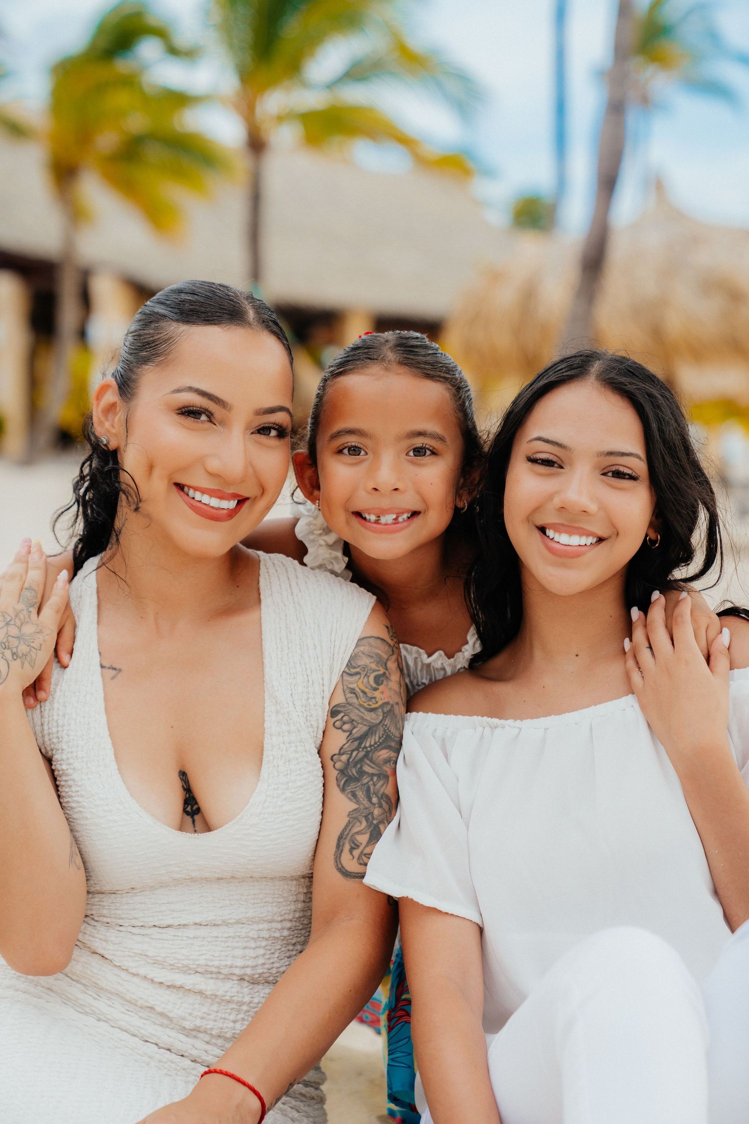 Family members in white outfits share joyful moments on a tropical beach with palm trees in the background.