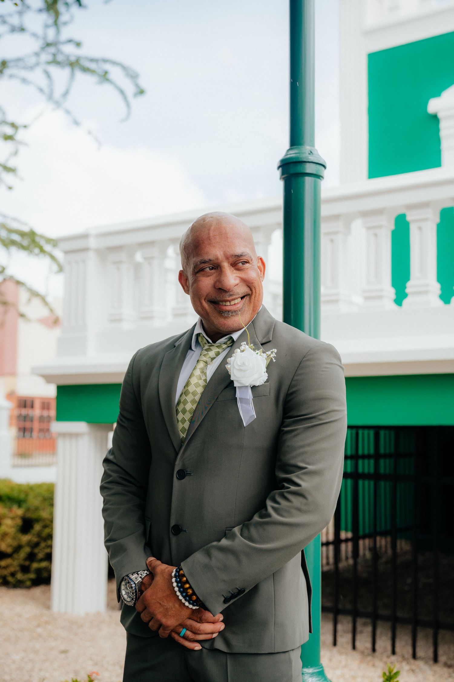 Person in dark suit with boutonniere smiles warmly outside green building.
