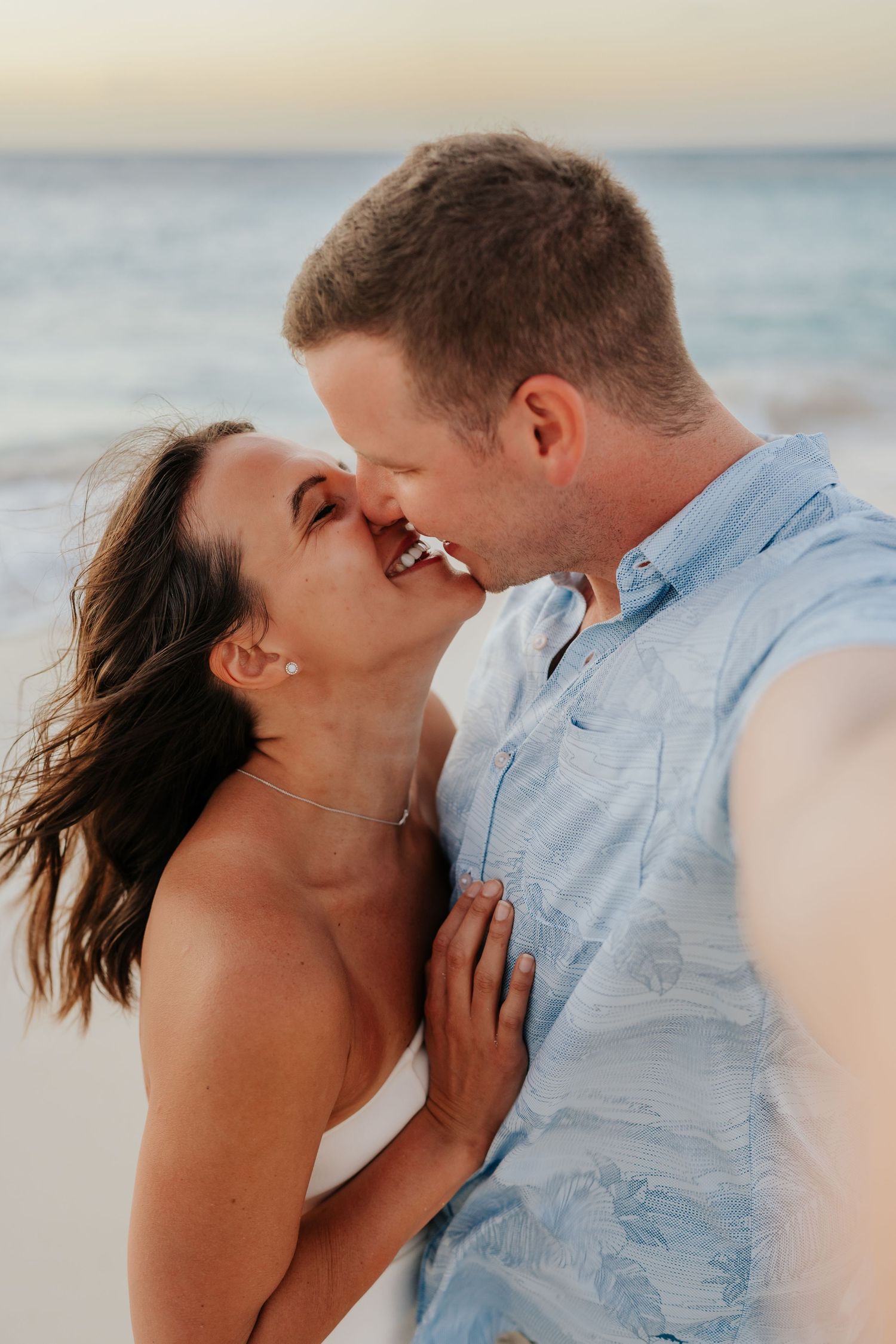 A couple shares a joyful almost-kiss on a beach at sunset, the woman laughing as waves crash behind them.