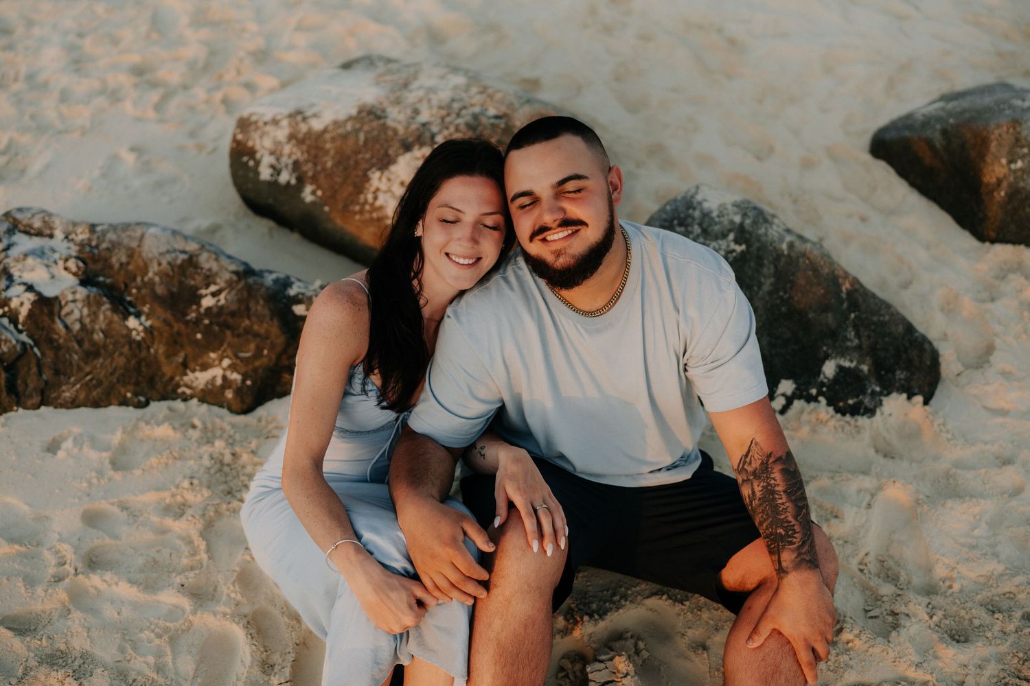 A smiling couple sits together on a sandy beach near rocks, the man with a tattoo wearing a white t-shirt.