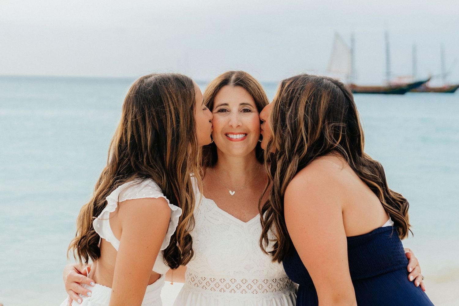 Three people share an intimate moment on the beach with sailboats visible in the distance.