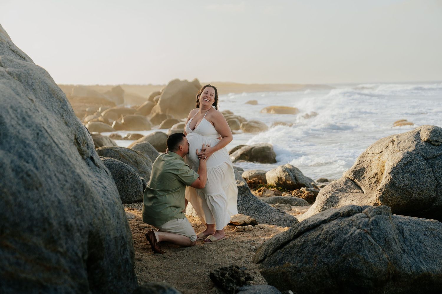 Couple shares a tender moment on rocky beach at sunset, pregnant woman in white dress with partner seated on sand.