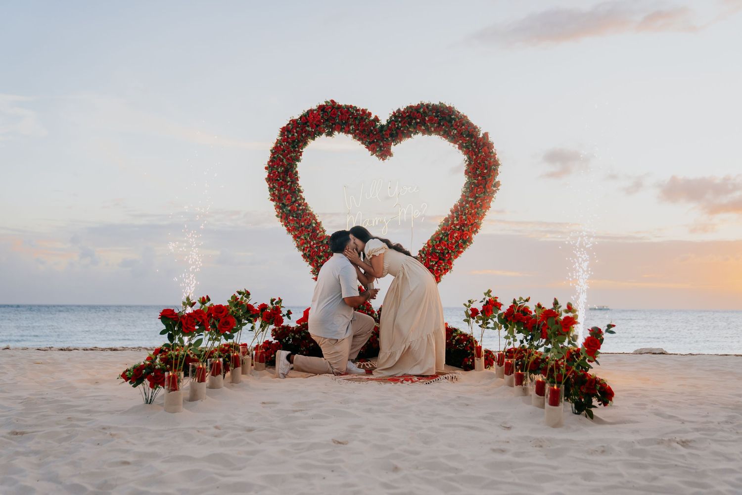 Romantic beach proposal with a large red rose heart arch, candles, and flowers at sunset on white sand.