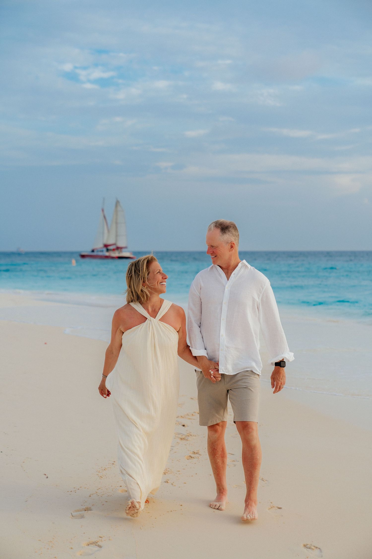 A couple takes a romantic beach walk with a sailboat visible on the turquoise horizon.